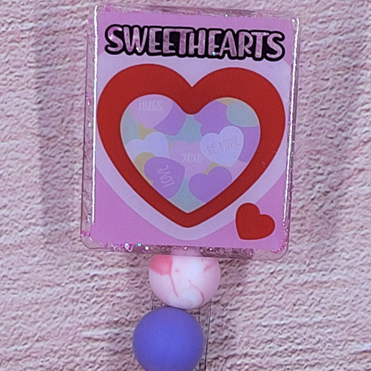 Experience the iconic Valentine's Day candy in a whole new way with our Badge Reel Candy Hearts. Featuring a pink shimmering base and coordinated beads, this is the perfect accessory for the holiday season.