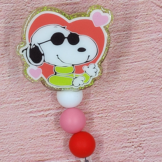 The Badge Reel Valentine Love Hound features Snoopy, the lovable pooch, ready for Valentine's day. Cool shades and hearts are highlighted on a green glitter base with matching beads in the background