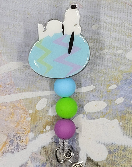 Everyone's Favorite Beagle has replaced is dog house with an Easter Egg as he takes his daily nap. Laying on a light blue egg with pink and green zig zag stripes on a clear glitter background and adorned with three perfectly matching silicone beads, adding a charming touch to this delightful accessory. A must-have for all animal enthusiasts!