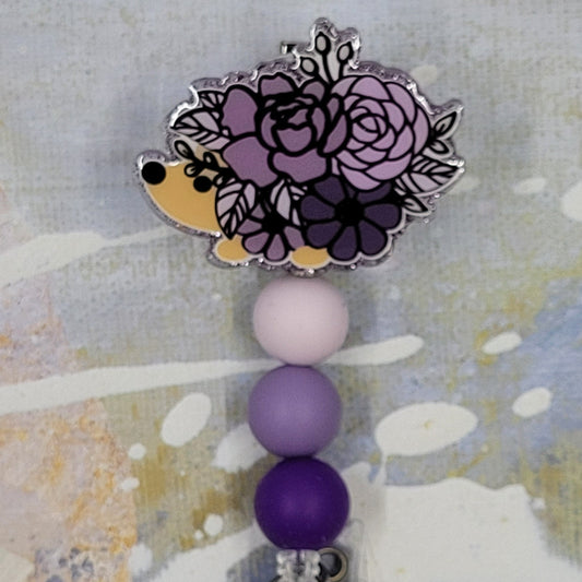 This Badge Reel features a cute purple floral hedgehog design paired with a shiny purple glitter background. The adorable hedgehog is complemented by three color-coordinated silicone beads for an extra touch of charm. Sure to be a hit with any animal lover.