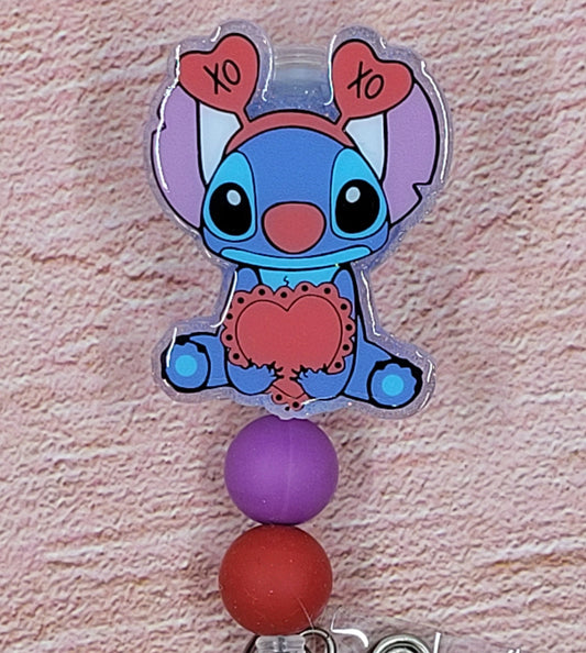 This badge reel features Experiment 626 asking you to be his Valentine. Wearing a cute head band with hearts and holding a big heart, this cute lil feller is adorable. A blue glitter base and 3 color coordinated beads finish the look..