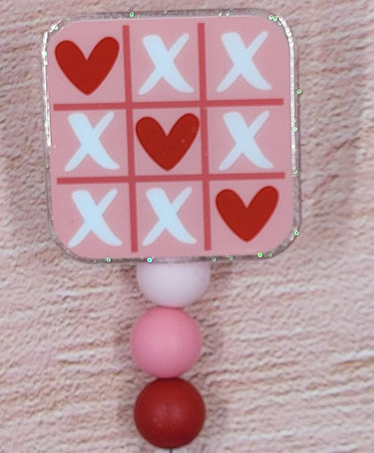 Tic Tac Toe has never been this cute where hearts are the big winner. The classic game with x's and hearts in white and reds on the pink base board Adding to its festive appeal is an iridescent glitter base, accompanied by three silicone beads in coordinated colors. Elevating your style for the holiday season, this is a must-have accessory.