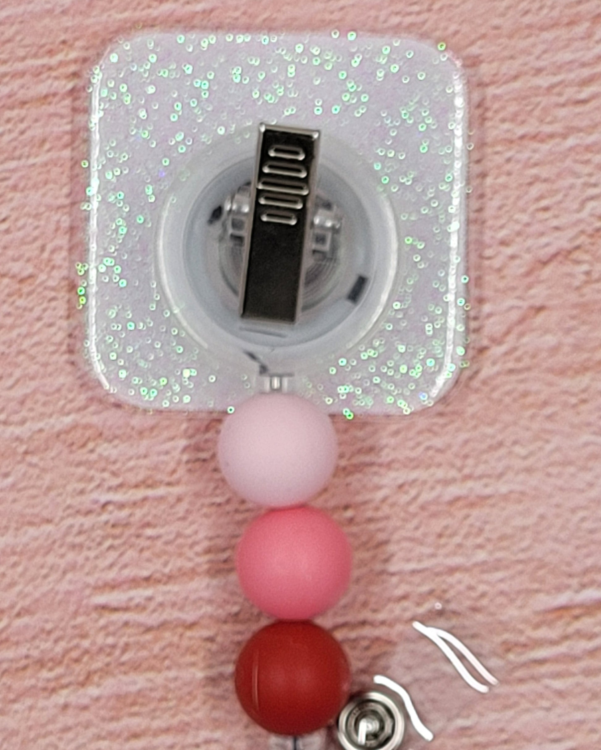 Tic Tac Toe has never been this cute where hearts are the big winner. The classic game with x's and hearts in white and reds on the pink base board Adding to its festive appeal is an iridescent glitter base, accompanied by three silicone beads in coordinated colors. Elevating your style for the holiday season, this is a must-have accessory.