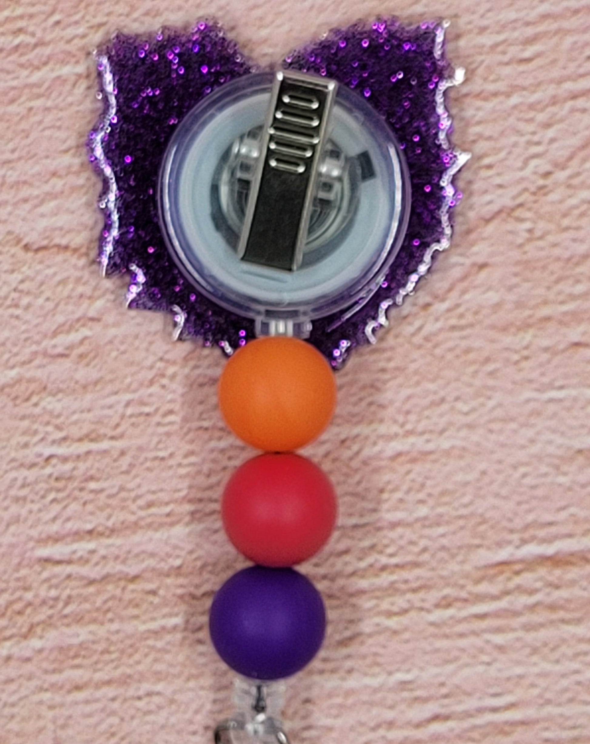 This Badge Reel utilizes an attractive floral pattern in a bold color palette. The design is complemented by a purple glitter base and three coordinated silicone beads.