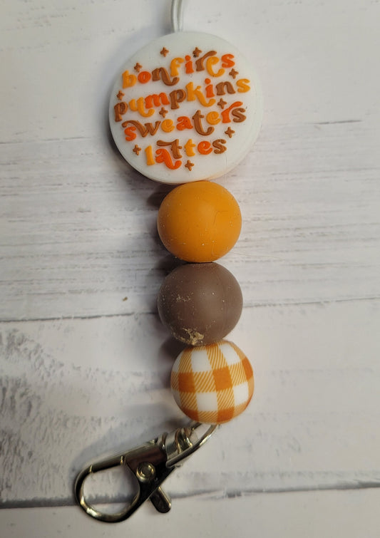 Need a custom Freshie Hanger? Here is a silicone beaded hanger we like to call Fall Vibes. All silicone bead construction with a sturdy clip to hang your Freshie from. Features a round white focal bead with the words Bonfires Pumpkins Sweaters &amp; Lattes and color coordinating beads to finish it off. Attaches quickly to any one of our many Freshies so you can easily hang it from your rear view mirror.