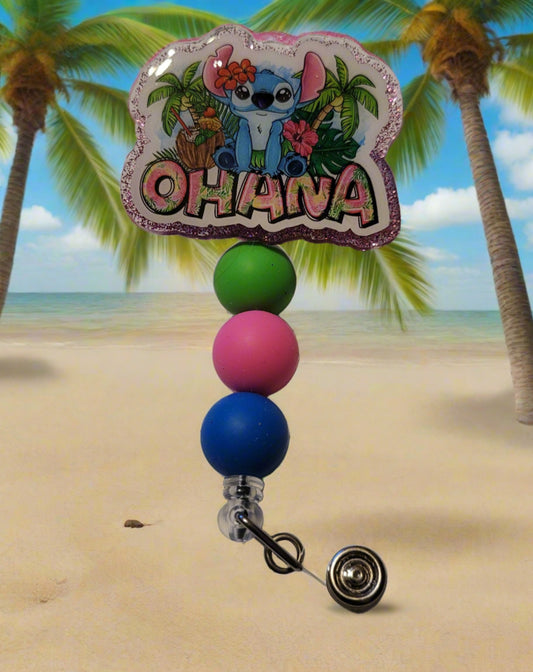 The Badge Reel Ohana showcases our happy little alien indulging in his preferred beverages on a palm tree covered beach. The elegant design is enhanced by a sparkling purplish glitter base and three color-coordinated beads.