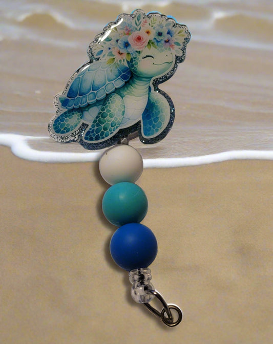 How cute is this happy little blue turtle with a floral head band! A beautiful bouquet sits on his head with a smile on his face. Features a blue glitter back and an adorable trio of matching silicone beads for a dash of extra charm. Perfect for all animal enthusiasts!