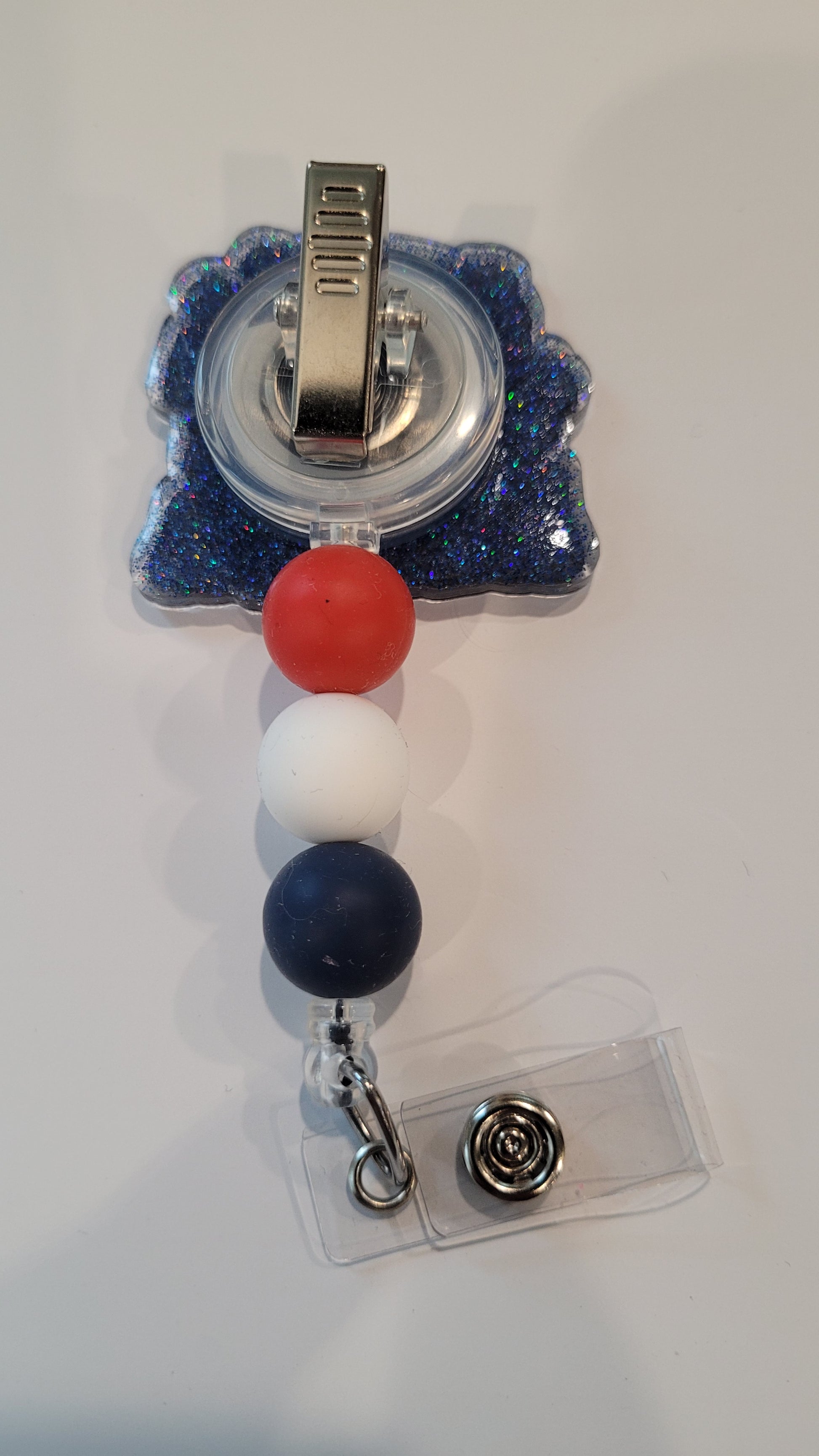 Experience America like never before with the Badge Reel America Alien Style! Embrace the summer spirit with cookouts, fireworks, ice cream, and everyone's favorite little blue alien. A dazzling blue glitter backing and 3 color-coordinated silicone beads add the perfect finishing touch to this out-of-this-world badge reel.