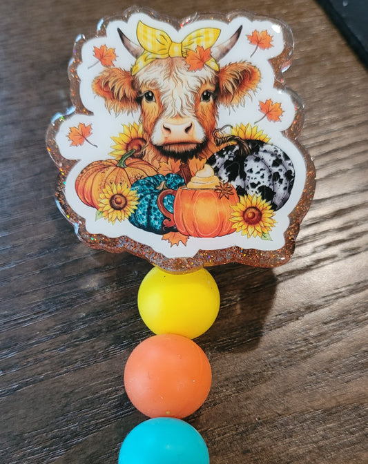 The classic Highland Cow is here in a fall harvest theme. With a yellow ribbon on her head she is surrounded by pumpkins and maple leaves in the traditional fall like feel. An orange glitter back and 3 silicone beads finish the look.