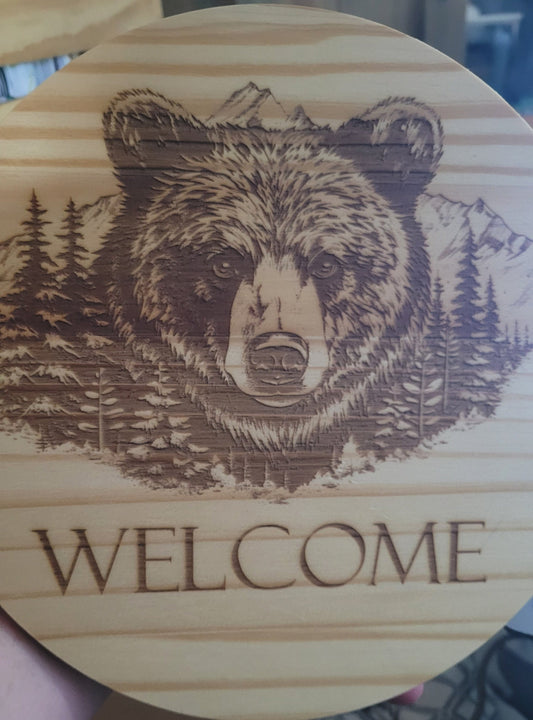 Here we took a 5/8" thick x 10" round pine board and laser engraved a Bear with a mountain range in the background. Also has they word "Welcome" under the main image. Sealed with multiple coats of clear poly to help protect the finish. 1 sawtooth and 2 nails included for hanging.