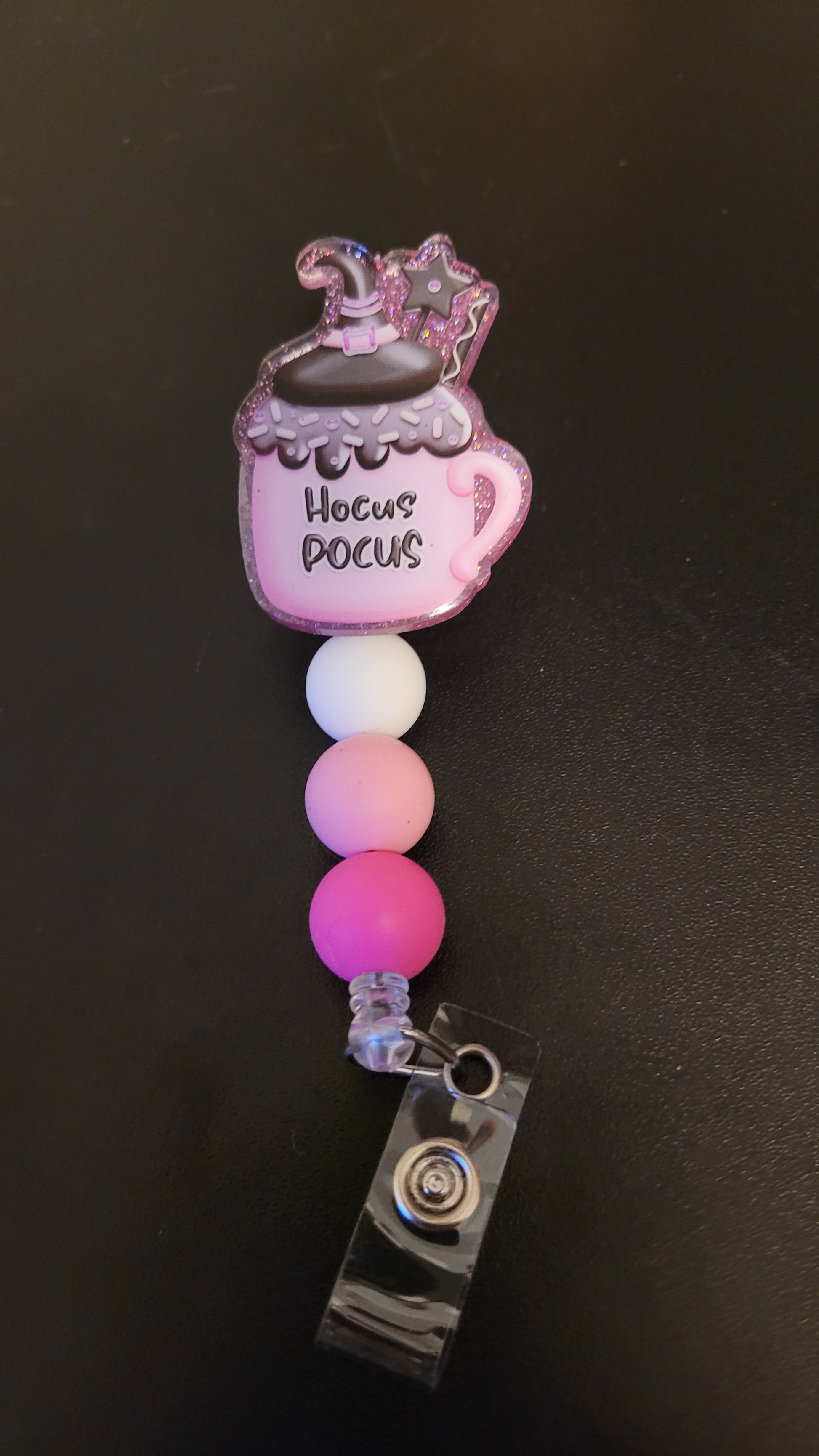 This sleek acrylic badge reel features a black witch hat and wand, perched atop a pink mug with Hocus Pocus in black text containing a warm cup of hot chocolate. The design is complete with a white silicone bead and two varied shades of pink.