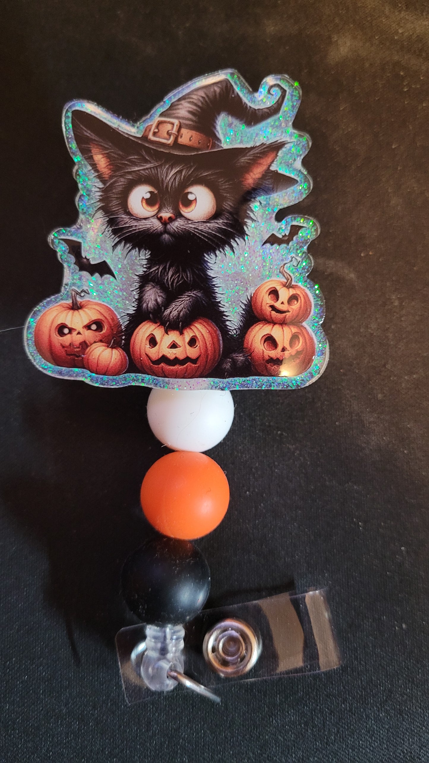 This pumpkin acrylic badge reel features a frazzled black witch cat planning his next scary trick.... or treat. A shiny glitter base pops through and is finished with 3 color coordinated silicone beads.