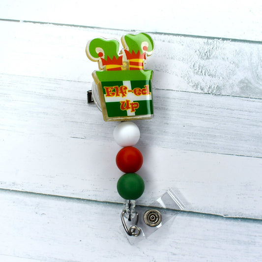 This acrylic badge reel features elf feet emerging from a brick chimney in vibrant white, red, and green, creating a visually-appealing Christmas theme.