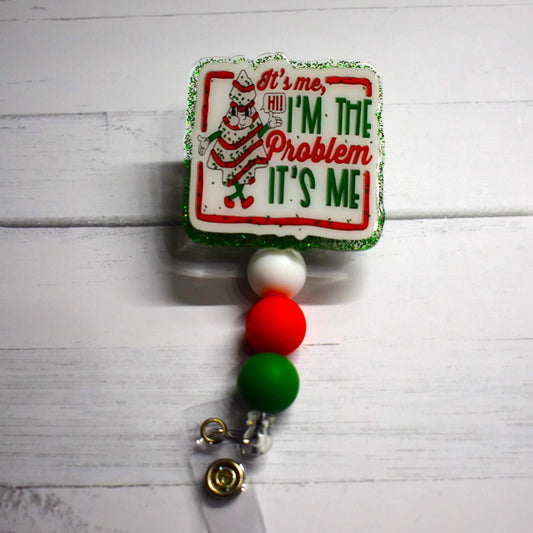 This acrylic badge reel proudly displays a classic holiday treat - the Christmas Tree Snack cake and he is the problem! A white background over a green glitter base and festive-hued silicone beads complete this seasonal delight, which is tastefully adorned with the words "It's Me, I'm The Problem It's Me".