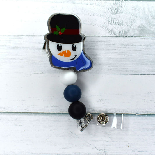 As snowflakes drift to the ground, a snowman appears with his iconic top hat and carrot nose. A navy, white, and black combination of 3 silicone beads complete the look of this acrylic badge reel.