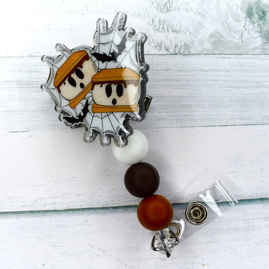 Creepy Camping out often leads to enjoying the classic treat of smores! This attractive acrylic badge reel offers a scary depiction of 2 smores on a spiderweb background, complete with white chocolate-brown & light brown silicone beads.