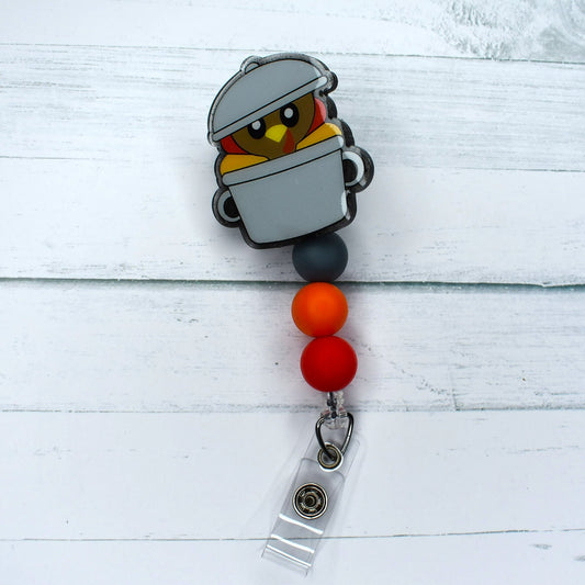 Thanksgiving Turkey peaking out of a roasting pan is the theme of this acrylic badge reel featuring grey, orange, and red accent silicone beads.