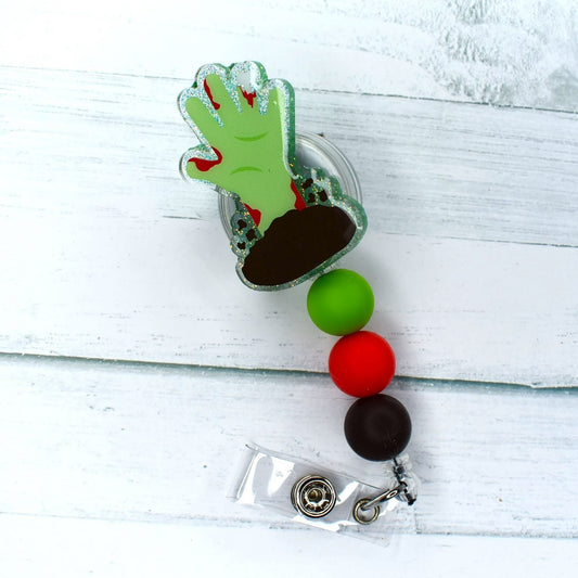 An undead arm rises from the soil, featured on this acrylic badge reel designed to evoke the feeling of a post-apocalyptic world. The accessory is finished off with green, red, and brown silicone beads.