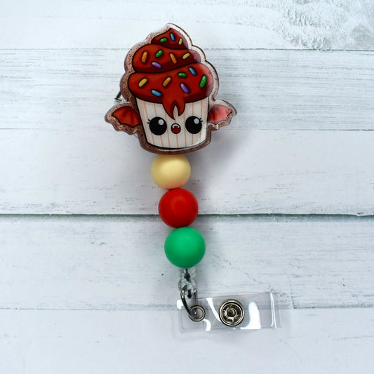 This acrylic badge reel boasts a delightful cupcake bat adorned with red frosting and sprinkles, perfectly complemented by cream red and green silicone beads.