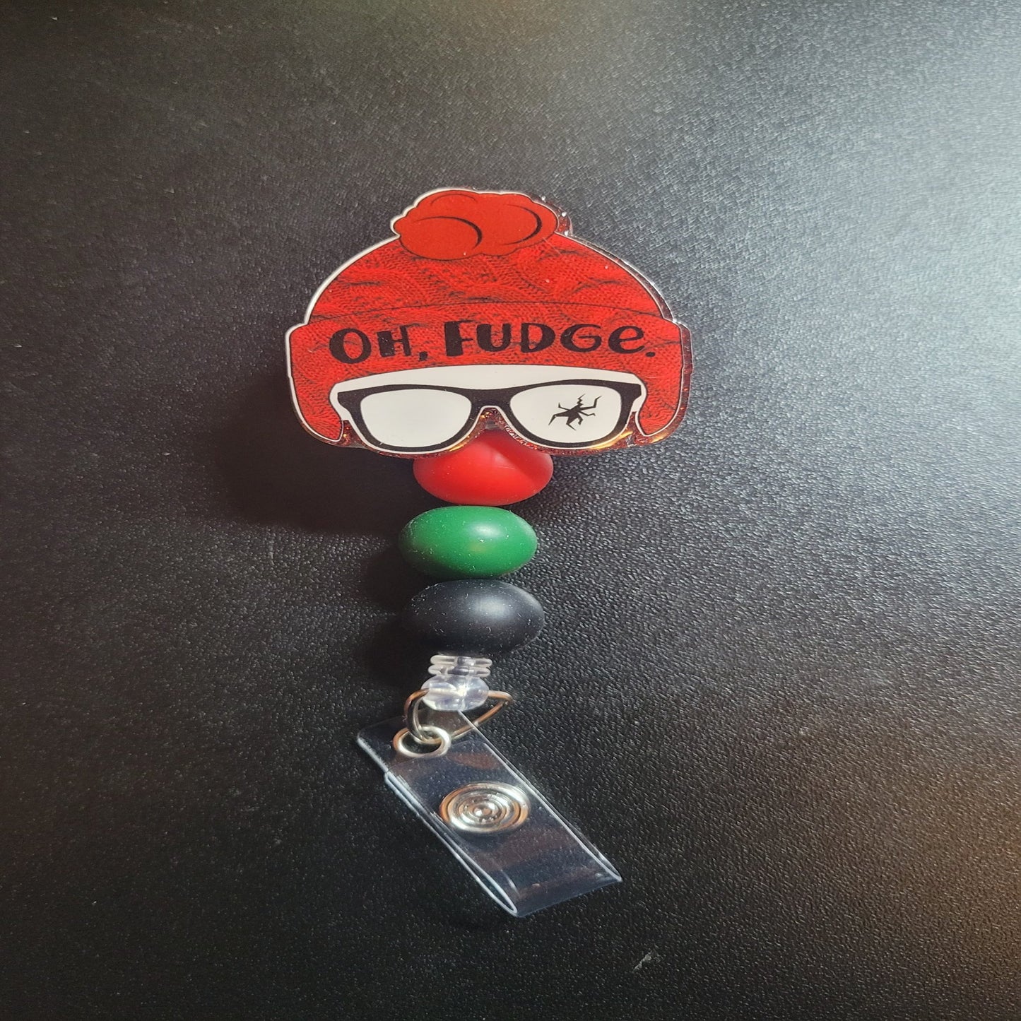 This acrylic badge reel prominently displays Ralphie from the beloved Christmas Story movie. Characterized by a cracked glasses lens and two words--Oh Fudge--this item is accented by three silicone beads in green, red, and black.