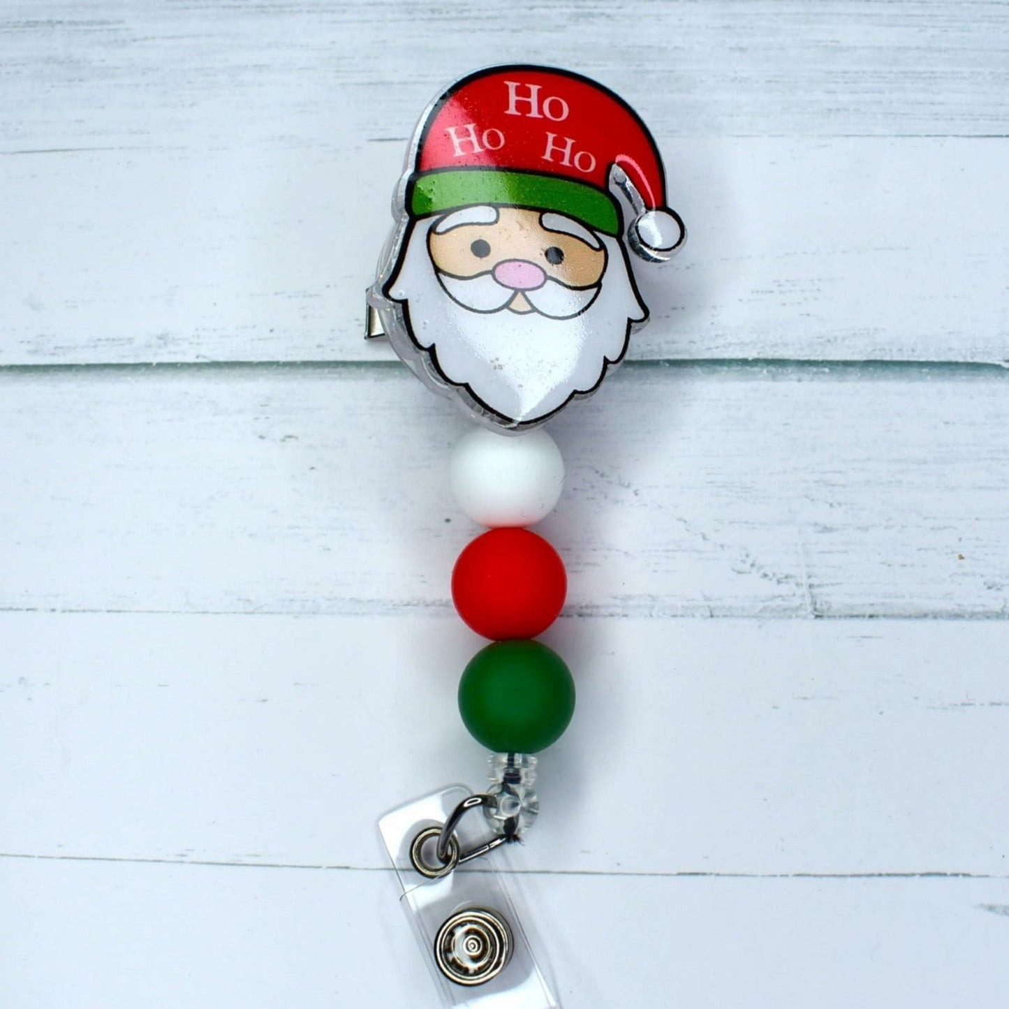 This acrylic Santa badge reel provides festive cheer with its design featuring "Ho Ho Ho" on the stocking cap, along with white, green, and red silicone beads. An excellent choice for a stocking stuffer.