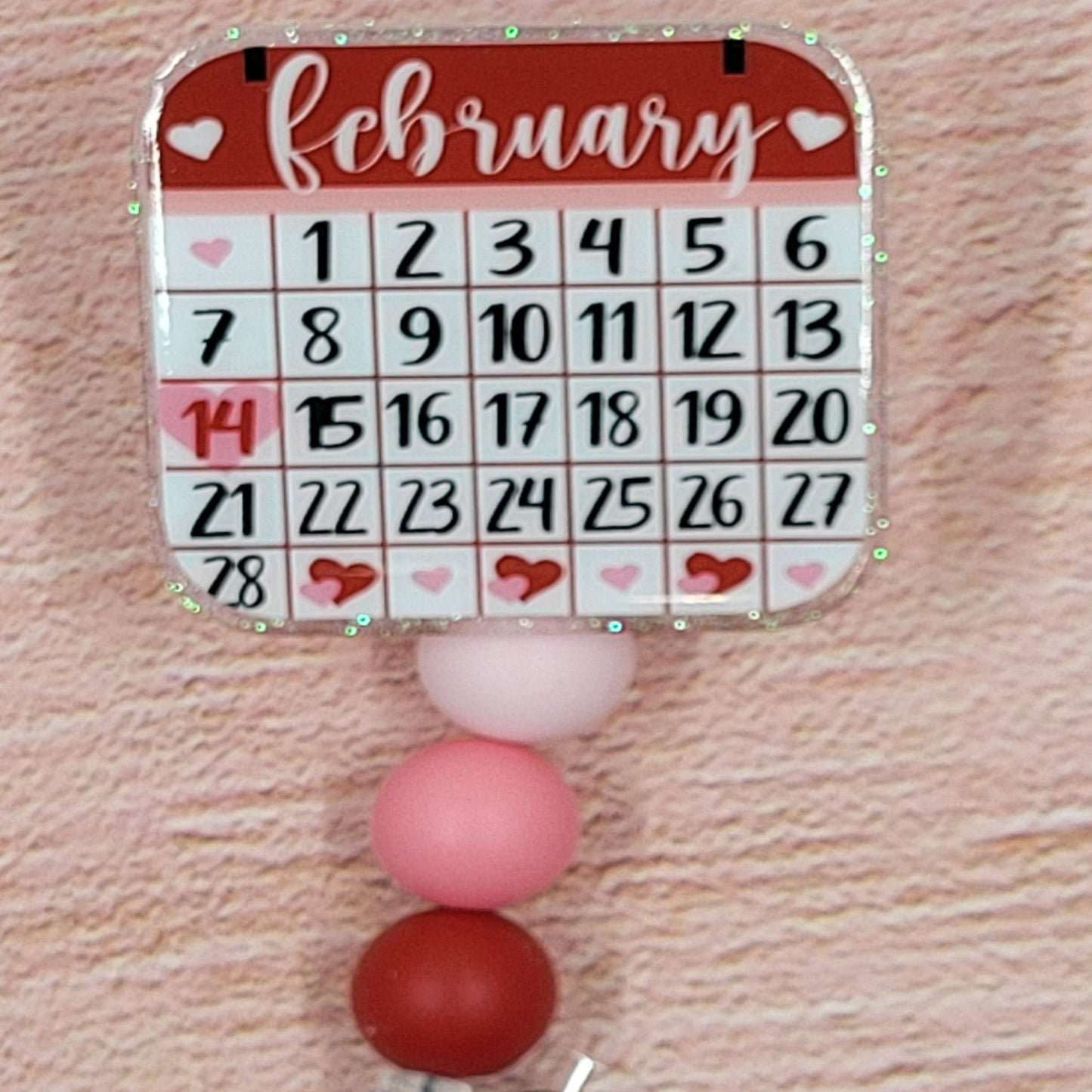 This 2" x 2" Badge Reel features a Valentines themed February Calendar in a combination of pink and red hues. Adding to its festive appeal is an iridescent glitter base, accompanied by three silicone beads in coordinated colors. Elevating your style for the holiday season, this is a must-have accessory.