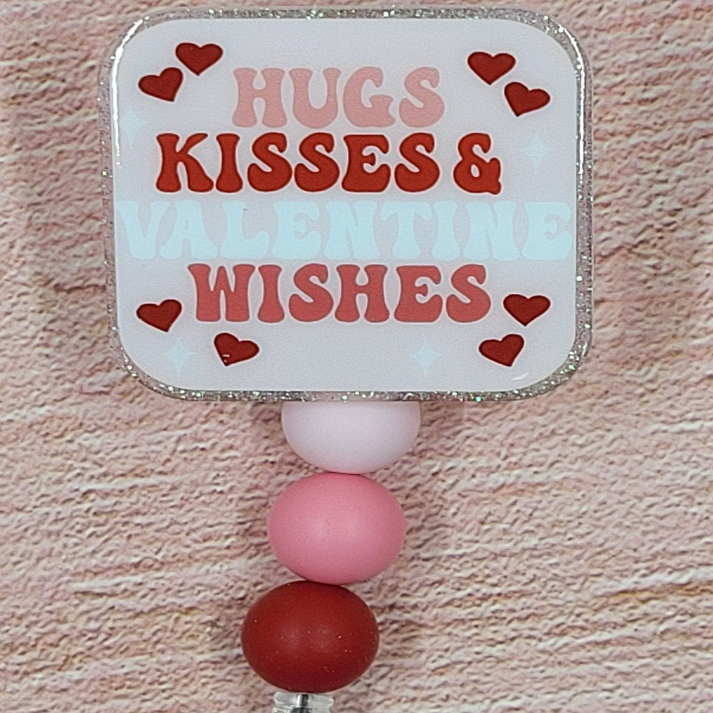 This 2" x 2" Badge Reel features a delightful combination of pink and red hues, adorned with the charming phrase "Hugs Kisses &amp; Valentine Wishes". Adding to its festive appeal is an iridescent glitter base, accompanied by three silicone beads in coordinated colors. Elevating your style for the holiday season, this is a must-have accessory.