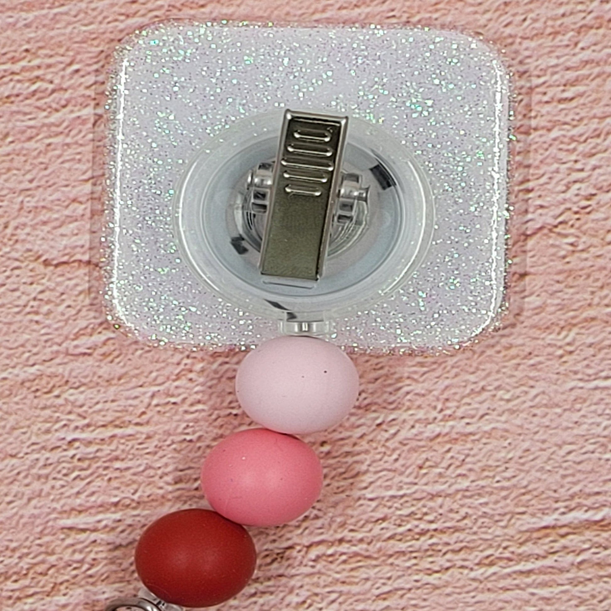 This 2" x 2" Badge Reel features a delightful combination of pink and red hues, adorned with the charming phrase "Hugs Kisses &amp; Valentine Wishes". Adding to its festive appeal is an iridescent glitter base, accompanied by three silicone beads in coordinated colors. Elevating your style for the holiday season, this is a must-have accessory.