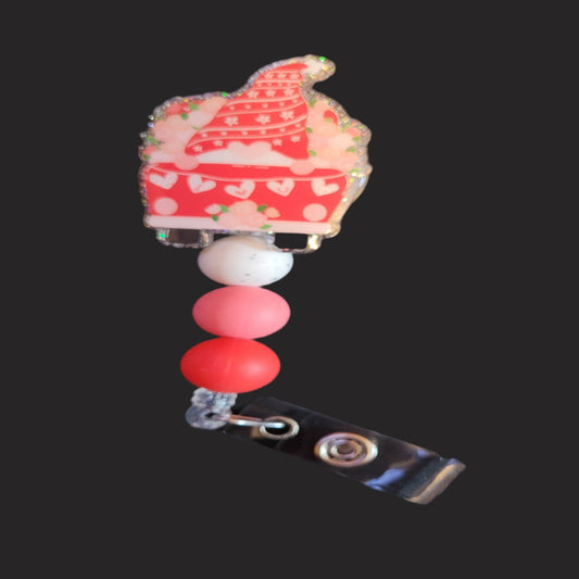 The Badge Reel Valentine Love Gnome Wagon features a cute gnome in a pink knit hat surrounded by flowers as he rides in a wagon on a mission to spread love. With a clear glitter base and 3 color matching silicone accent beads, this badge reel is sure to please.