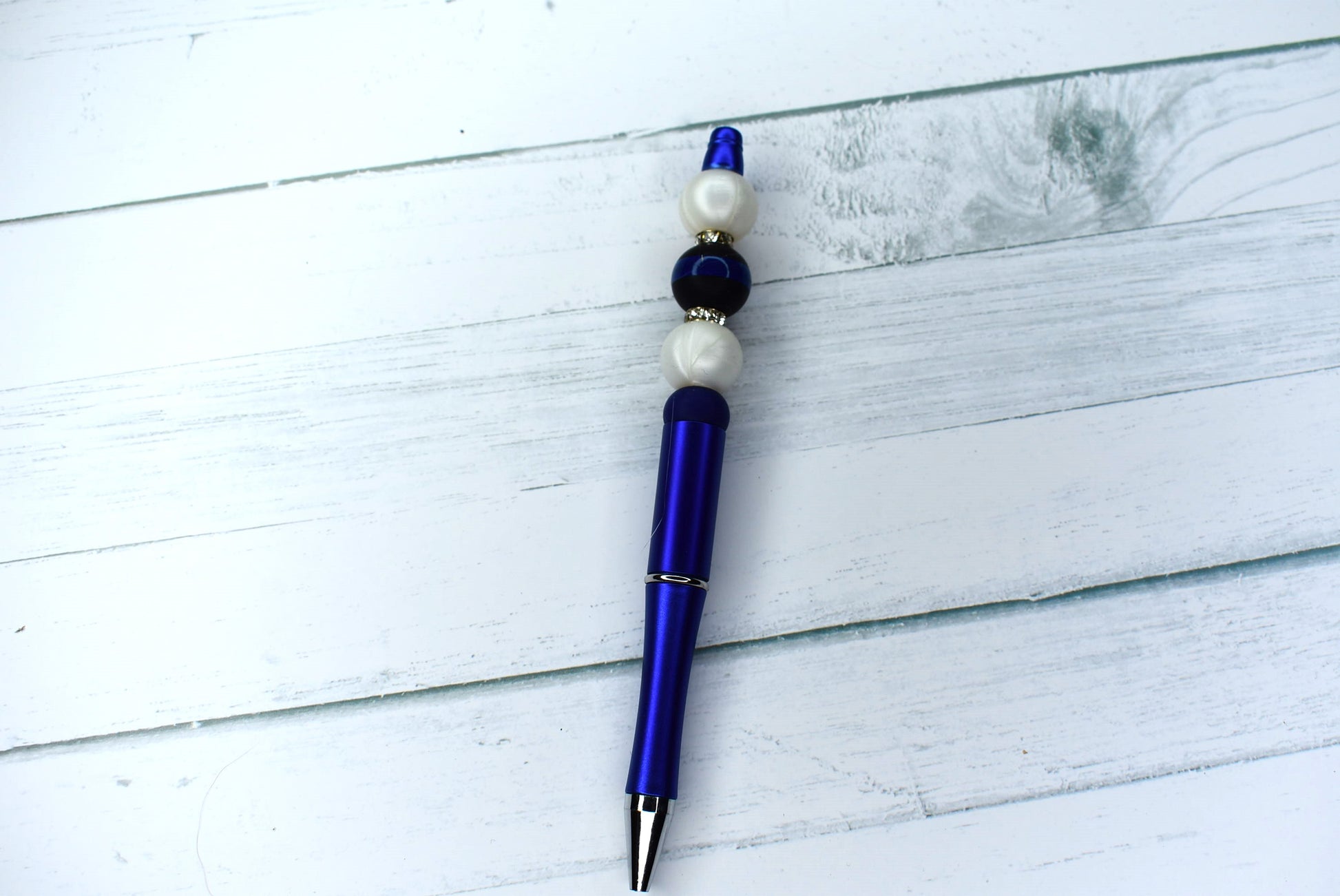 This striking pen features a blue base with a silicone black and blue striped focal bead. Sparkling gem rings and 2 pearl white beads finish the look. An extra black refill also included.