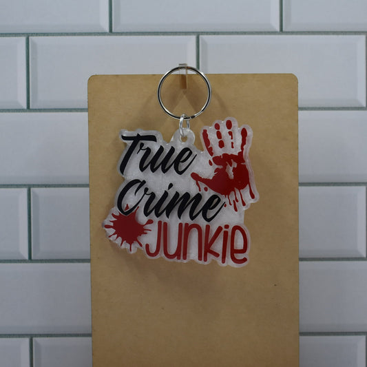  This acrylic keychain features a resin base with a silver powder coating and a bloody hand print accented with black and red detailing to signify the True Crime Junkie theme.