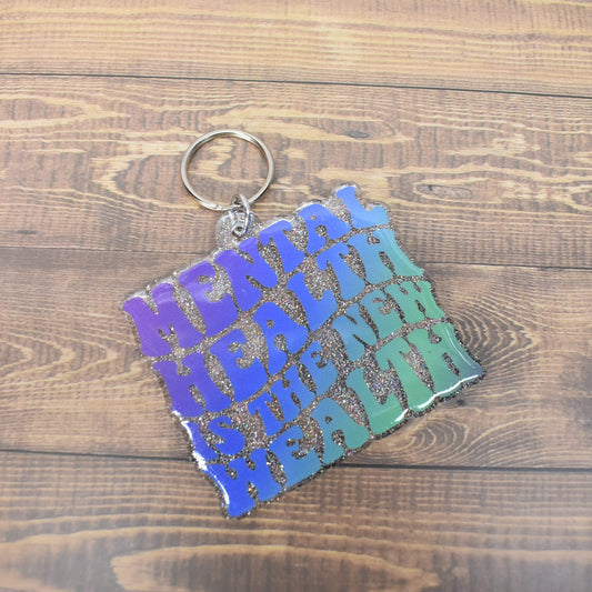 Acrylic keychain with the phrase Mental Health Is The New Wealth. Multi color that flows from purple to blue and ends in an Aqua Marine.